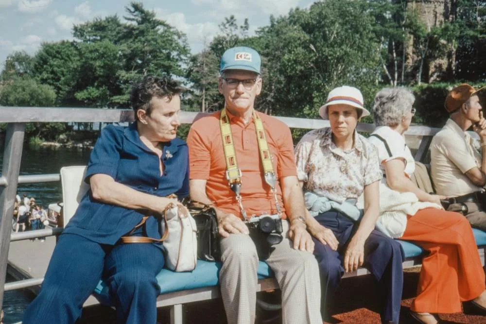 Older travelers sitting together on a bench beside a waterside railing, chatting and smiling