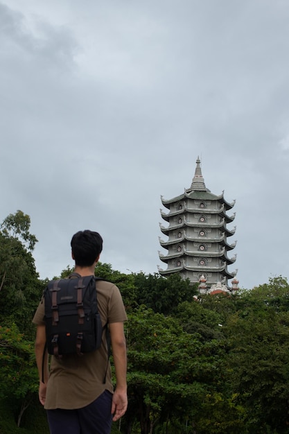 A traveler with a backpack looking up at a tall multi-tiered pagoda rising above green trees under a cloudy sky