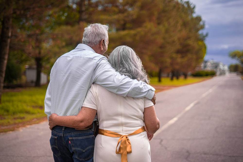 An older couple walking arm in arm along a quiet tree-lined road, seen from behind